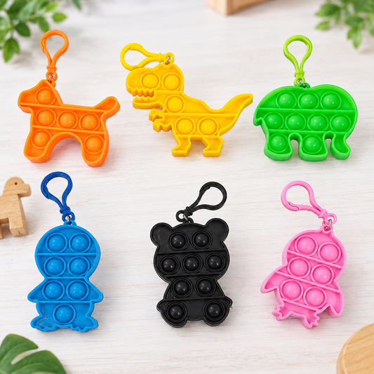 Six colorful fidget pop toy keychains shaped like a dog, dinosaur, elephant, turtle, bear, and chick, arranged on a light wood surface with green leaves in the background.