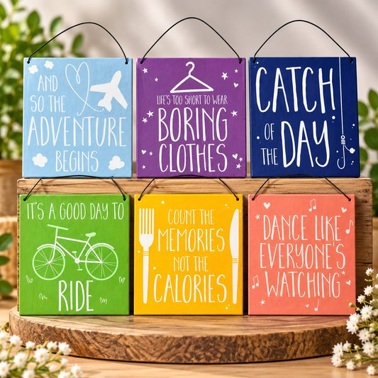 Six colourful hanging signs with black wire hangers are arranged on a round wooden tray. The signs feature playful white text and simple illustrations, including an airplane, hanger, fishhook, bicycle, fork, knife, and music notes.