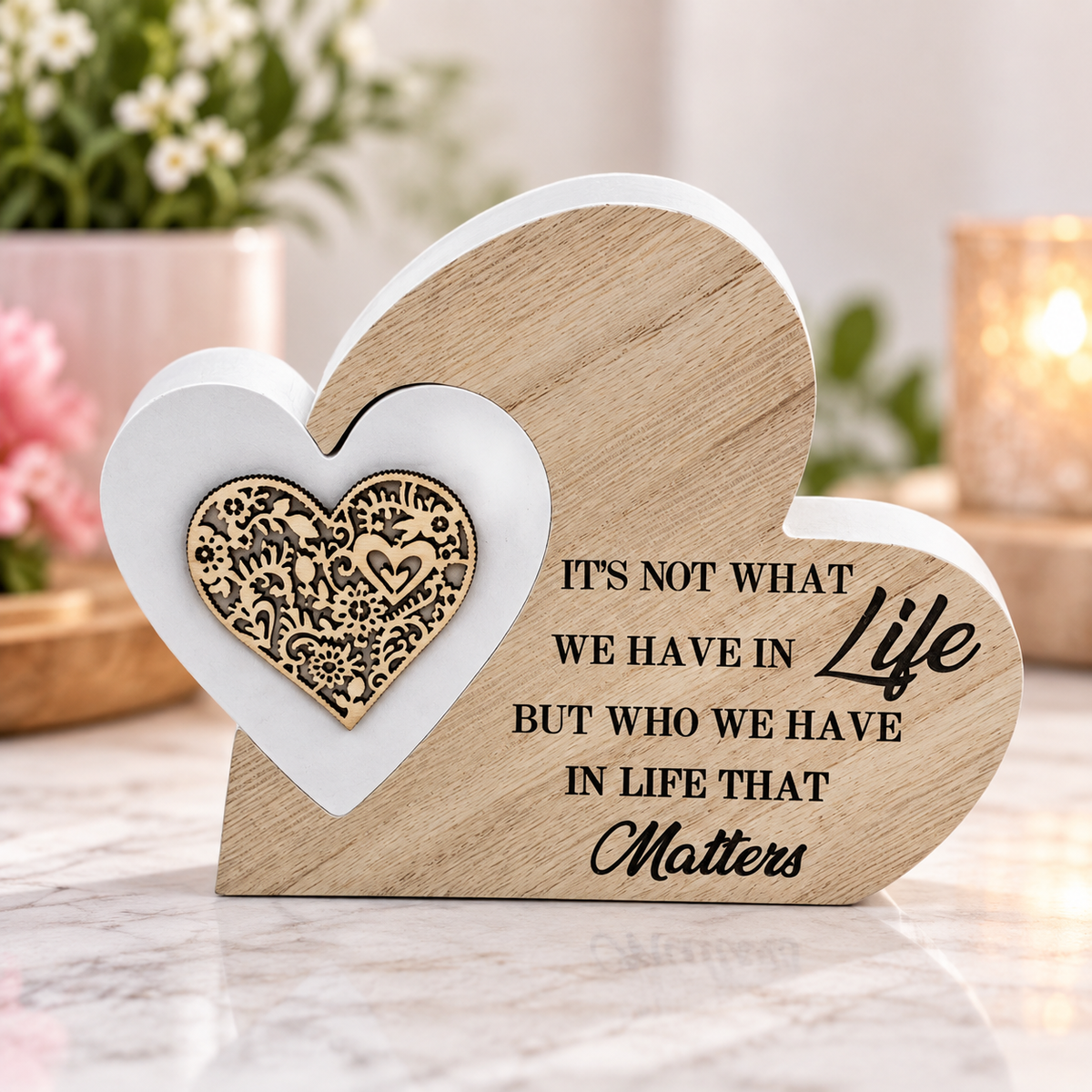 Heart-shaped wooden plaque with engraved text reading 'IT'S NOT WHAT WE HAVE IN Life BUT WHO WE HAVE IN LIFE THAT Matters' and a white heart with a gold ornate emblem, displayed on a marble surface with a blurred indoor background.