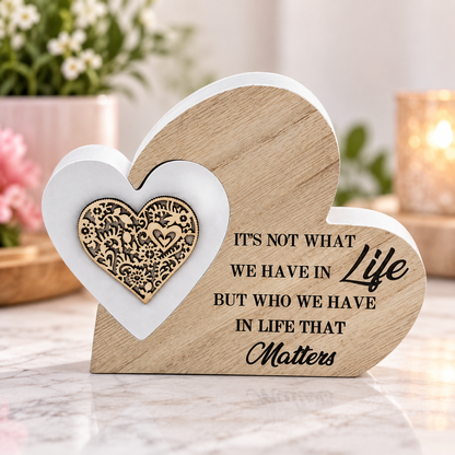 Heart-shaped wooden plaque with engraved text reading 'IT'S NOT WHAT WE HAVE IN Life BUT WHO WE HAVE IN LIFE THAT Matters' and a white heart with a gold ornate emblem, displayed on a marble surface with a blurred indoor background.