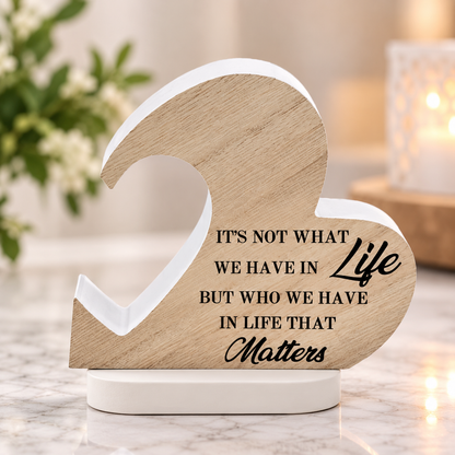A wooden heart-shaped tabletop sign with a white base, featuring the engraved quote, 'IT'S NOT WHAT WE HAVE IN Life BUT WHO WE HAVE IN LIFE THAT Matters'. The sign is styled on a marble surface with a blurred background containing a lit candle and greenery.