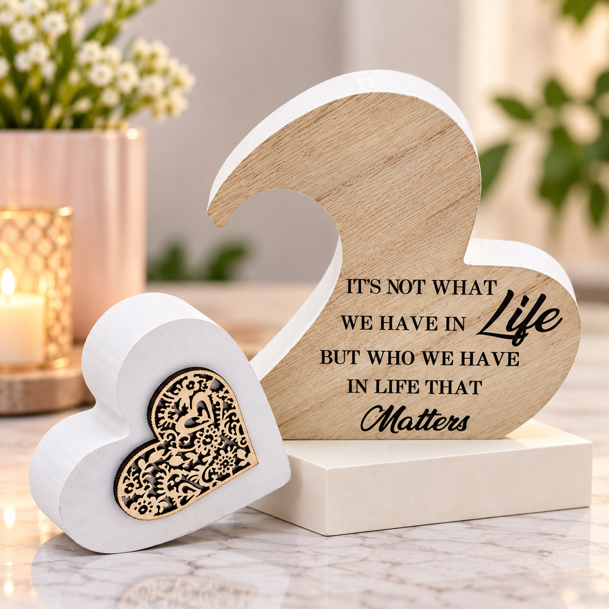 A decorative heart-shaped wooden and white memorial-style sculpture on a pedestal, with one heart showing engraved black text and the other featuring a gold-toned floral heart inlay, set on a marble surface with blurred flowers and candles in the background.