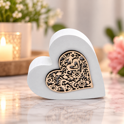 A white heart-shaped decorative block with a laser-cut black floral pattern on the front, displayed on a marble surface with a blurred candle and flowers in the background.