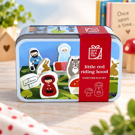 A children's storytime play set in a decorative tin box with a red wrap label reading 'little red riding hood'. The set includes illustrated character pieces and small accessories visible through the front of the tin, photographed on a tabletop with soft background props.
