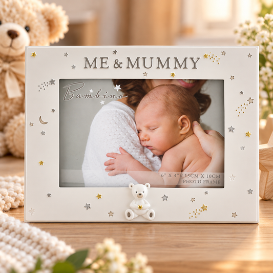 A personalized baby photo frame with 'ME & MUMMY' and 'Bambino' text, decorated with gold stars and a small white teddy bear at the bottom. The frame is shown on a wooden table with a blurred photo of a baby and mother inside, surrounded by a plush teddy bear and soft background decor.