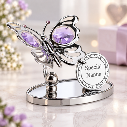 A silver butterfly-shaped keepsake holder with large purple faceted gemstones in its wings, mounted on a mirrored oval base. A round hanging tag reads 'Special Nanna.
