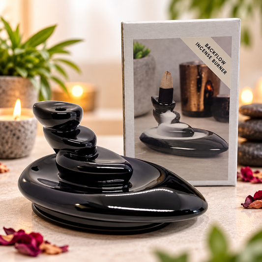 A glossy black back-flow incense burner with stacked rounded stones and a visible incense stick, shown in front of its retail box on a relaxing background.