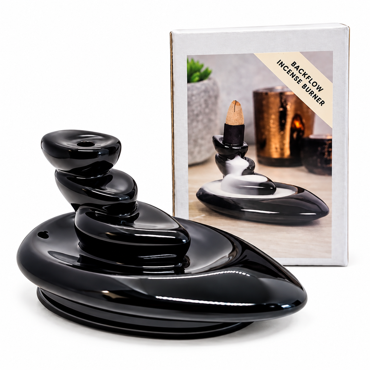 A glossy black back-flow incense burner with stacked rounded stones and a visible incense stick, shown in front of its retail box on a white background.