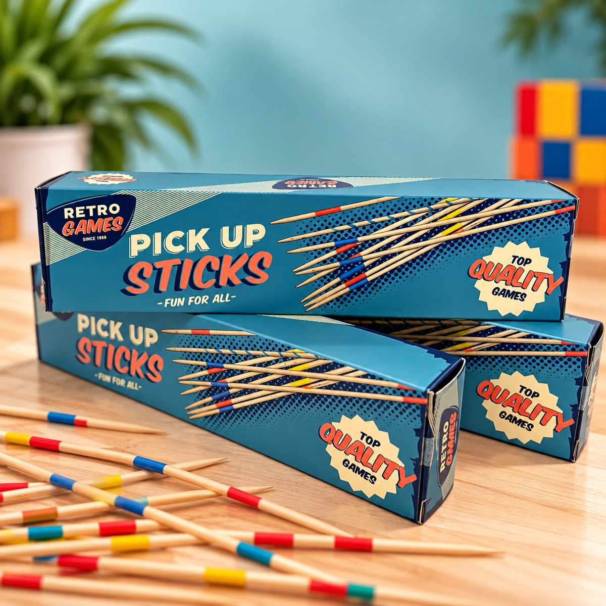 Three blue boxes of Retro Games Pick Up Sticks stacked on a wooden table, with several wooden sticks marked in red, yellow, and blue scattered in front. The background features a soft-focus blue wall, a green potted plant, and a colorful block toy.