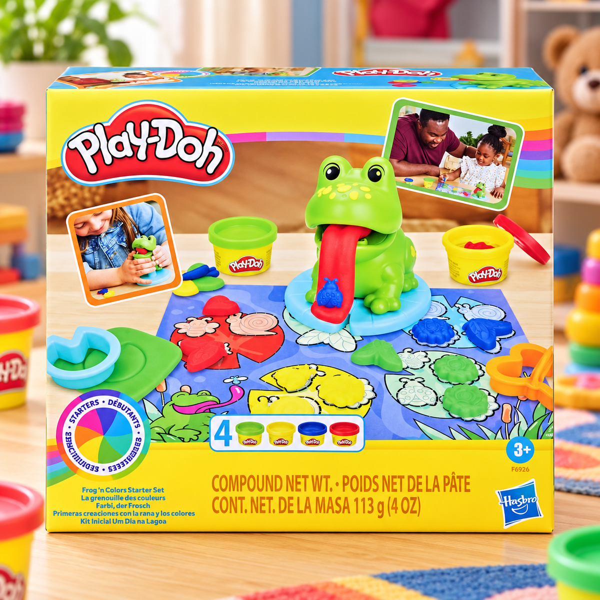 Play-Doh Compound Modeling Set box on a wooden table with colorful Play-Doh tools and clay bins arranged around it. The front of the box shows the Play-Doh logo, a green frog with a red tongue, and multiple color-changing clay shapes.