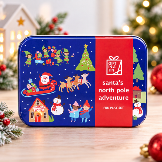 A rectangular metal tin with rounded corners labeled 'gift in a tin' and 'santa's north pole adventure FUN PLAY SET,' shown on a wooden surface with blurred Christmas decorations in the background. The tin has a dark blue left side with illustrated holiday scenes including Santa in a sleigh, elves, a Christmas tree, a snowman, and children, plus a red right side with white text.