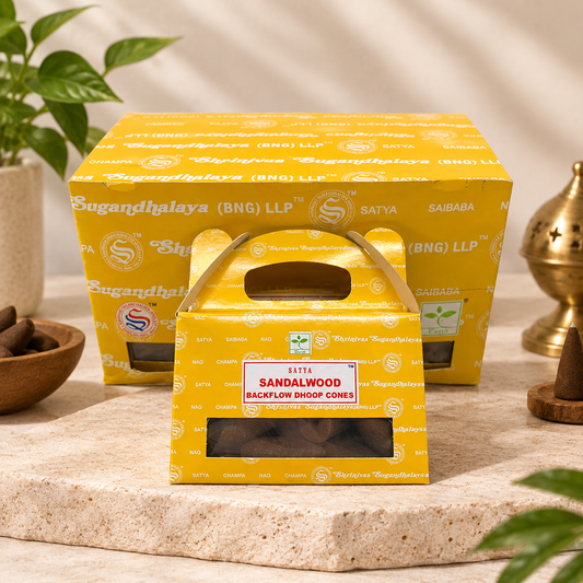 Yellow hand-carry boxes of Satya Sandalwood Backflow Dhoop Cones, with one box in the foreground and two stacked behind it, styled on a stone surface with a plant and brass incense burner in the background.