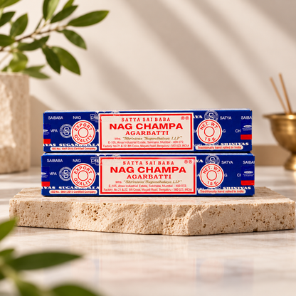 Two stacked boxes of Satya Sai Baba Nag Champa Agarbatti (incense sticks) on a stone slab with a blurred background showing a plant and a brass incense burner.