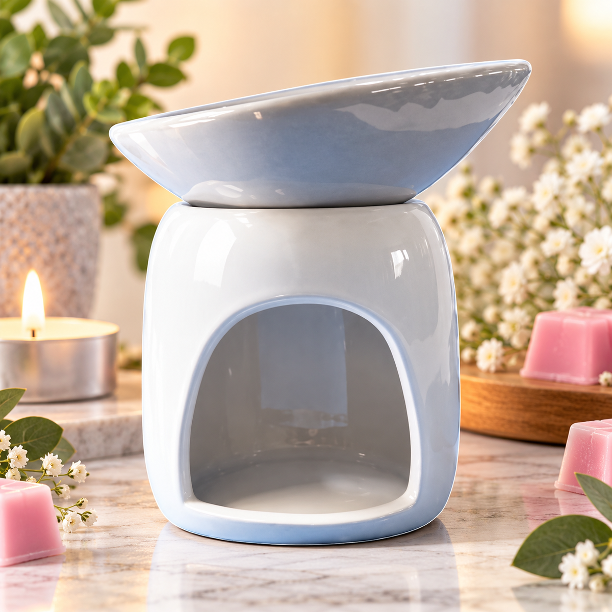 A ceramic wax warmer with a glossy light blue and white finish, styled on a marble surface with pink candles, white flowers, and a lit tea light candle in the background.