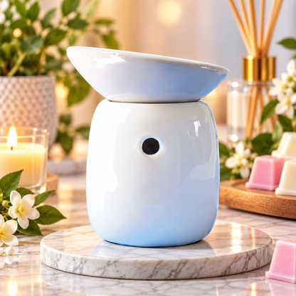 A ceramic oil burner warmer with a smooth white to light blue gradient finish, sitting on a white marble coaster. The background is a beautifully styled, warm-toned home setting with a lit candle, fresh flowers, and a reed diffuser, creating an inviting atmosphere.