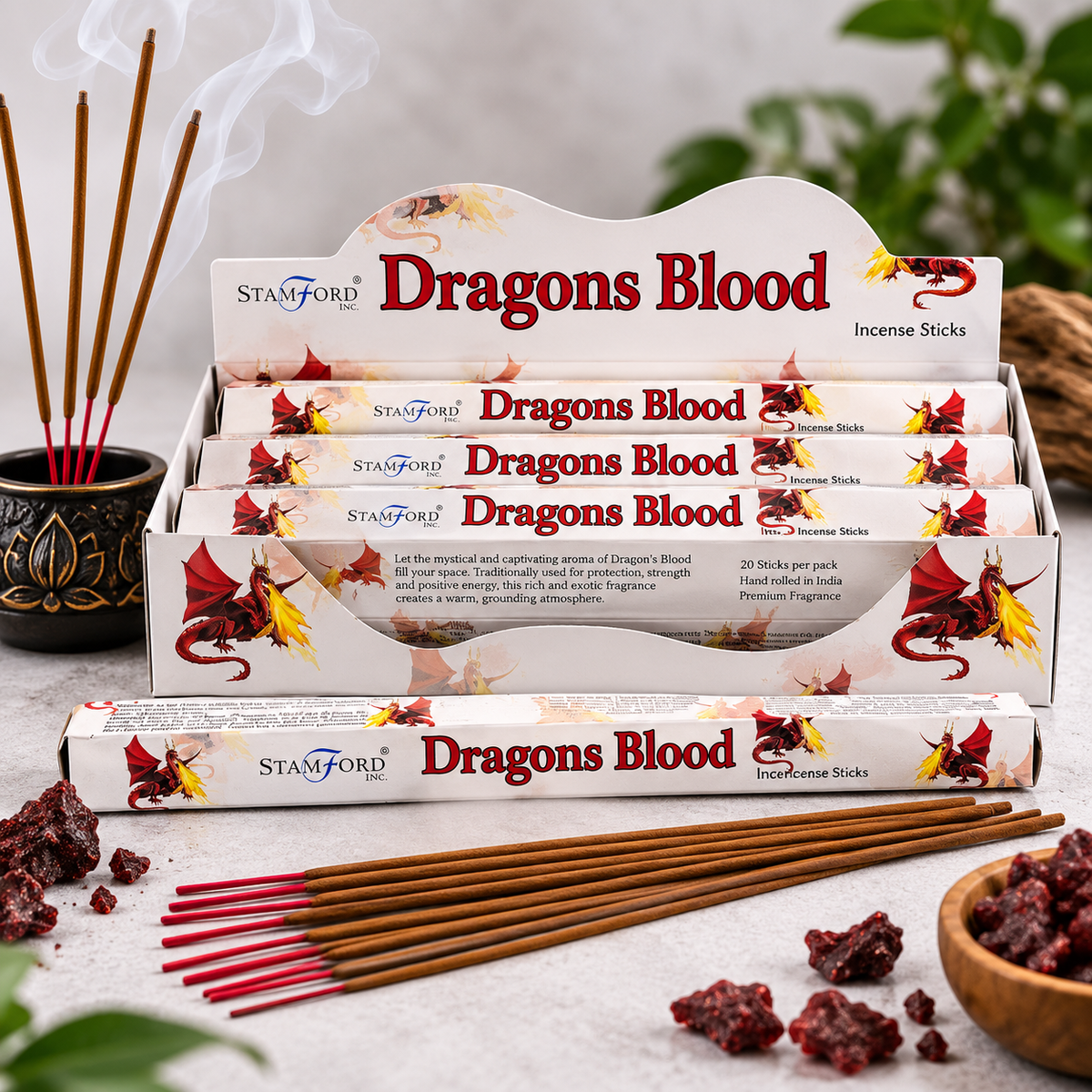 A display of Stamford Dragons Blood incense sticks, with several sticks burning in a black decorative pot on the left and loose incense pieces and a small bowl on the right, set on a light stone surface with soft green foliage in the background.
