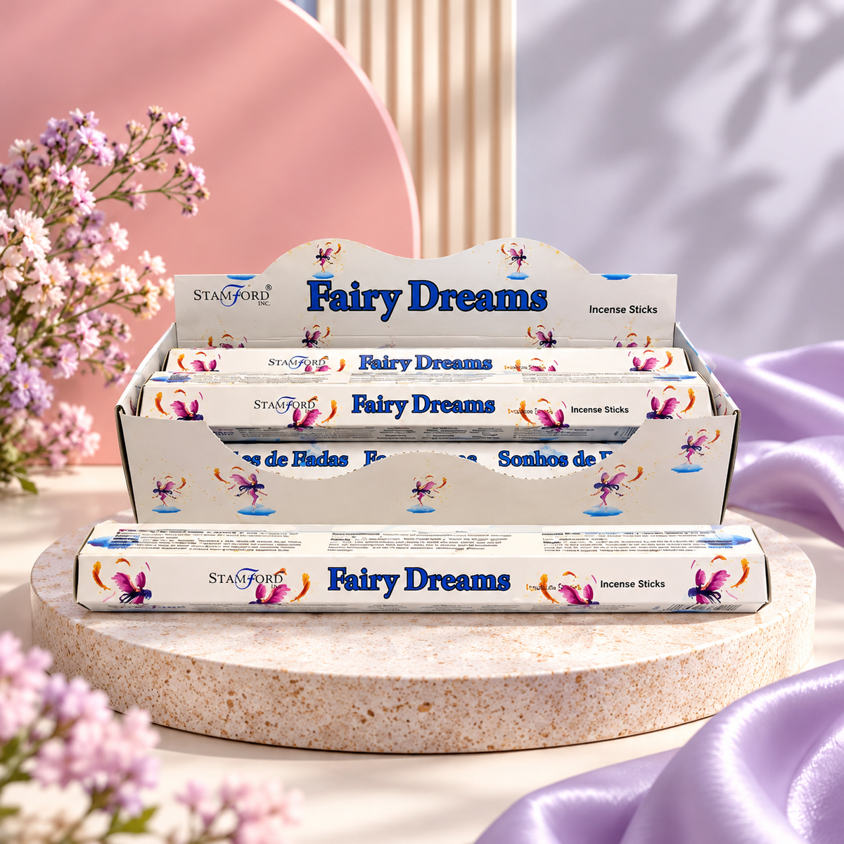 A display box and individual packs of Stanford Fairy Dreams incense sticks, styled on a round stone pedestal with soft pink and purple fabric and small flowers in the background.