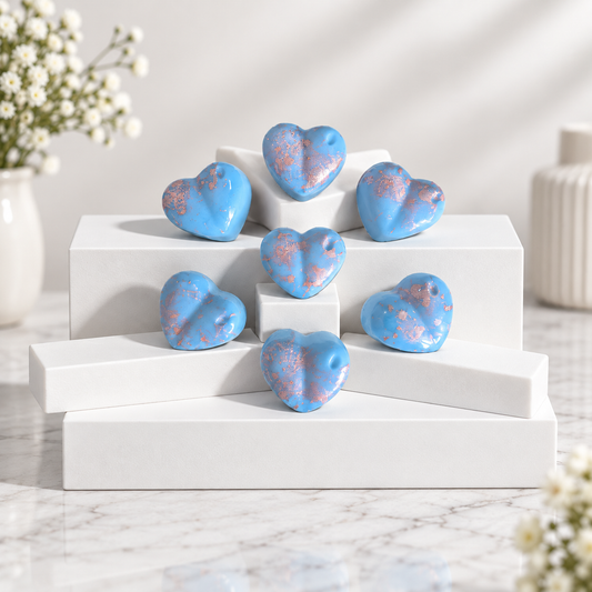 Six small heart-shaped Wax Melts in light blue with pink glitter accents, arranged on white display blocks over a marble surface with soft floral props in the background.
