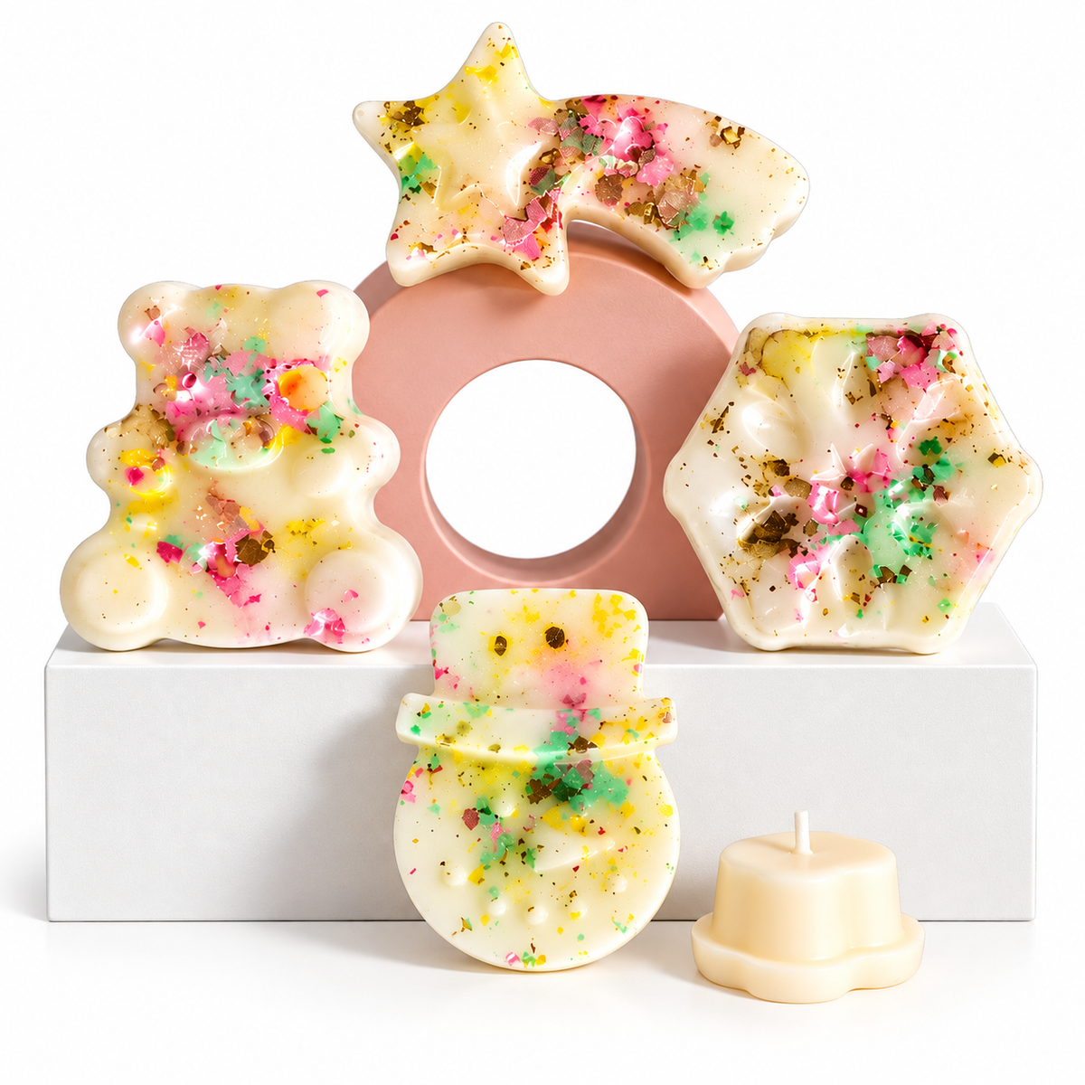 A set of decorative wax melts in playful shapes including a bear, star, snowman, hexagon, and a small taper candle, all in a creamy off-white base with colorful confetti and glitter flecks, arranged on a white block with a pink circular prop against a white background.