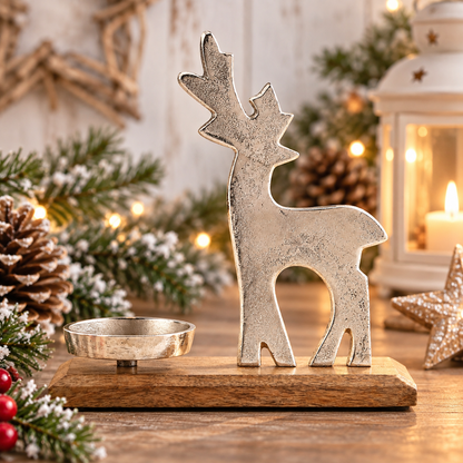 A silver-toned metal reindeer figurine with antlers stands on a rectangular wooden base next to a small matching metal bowl, styled on a rustic wooden table with pine branches, a lit lantern, and warm holiday lights in the background.