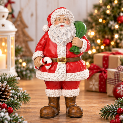 A Santa Claus figurine wearing a red coat with white trim, brown boots, and a gold belt, holding a green sack and a candy cane. The figurine is displayed on a wooden surface with a festive holiday background featuring a decorated Christmas tree, wrapped gifts, pine branches, and warm string lights.