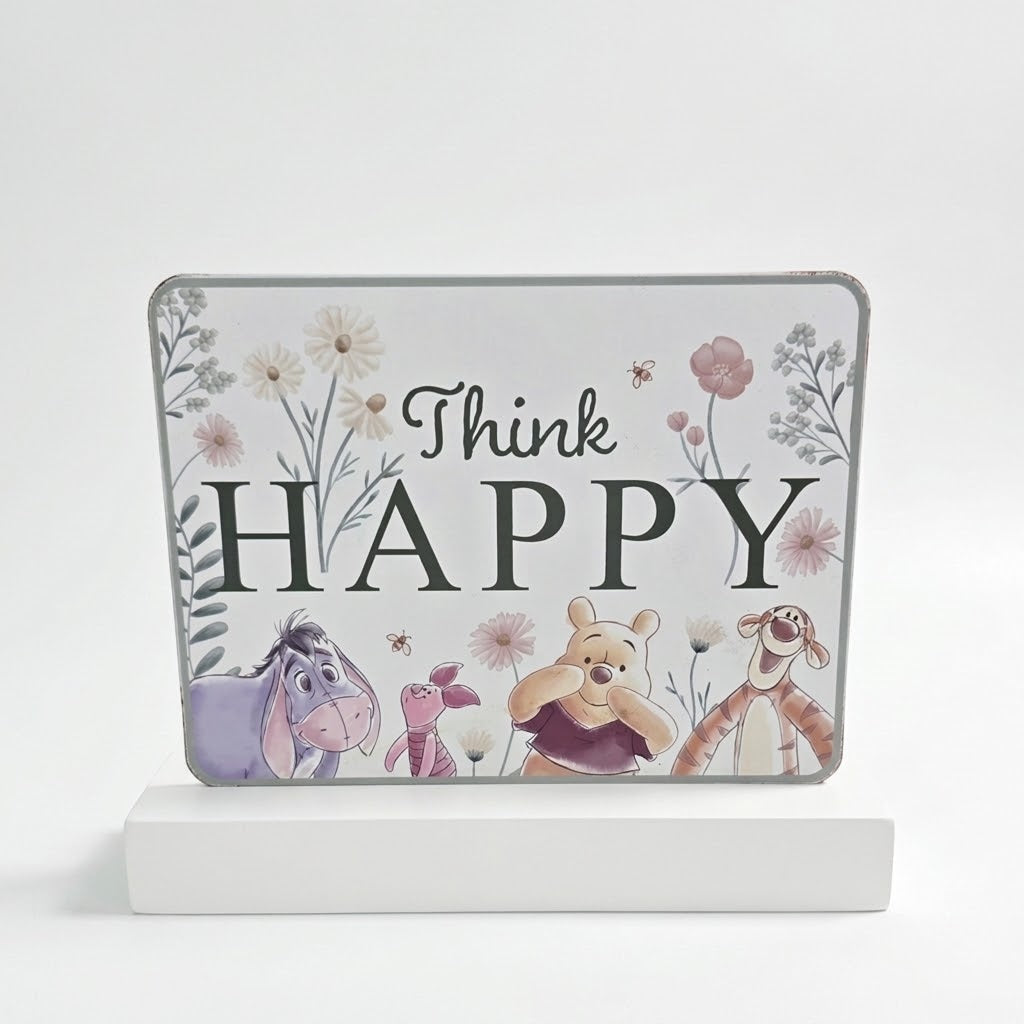 A decorative rectangular sign with rounded corners featuring the phrase 'Think HAPPY' in dark lettering, surrounded by pastel flowers and illustrated characters including Eeyore, Piglet, Winnie the Pooh, and Tigger. The sign is displayed upright on a white block against a clean white background.
