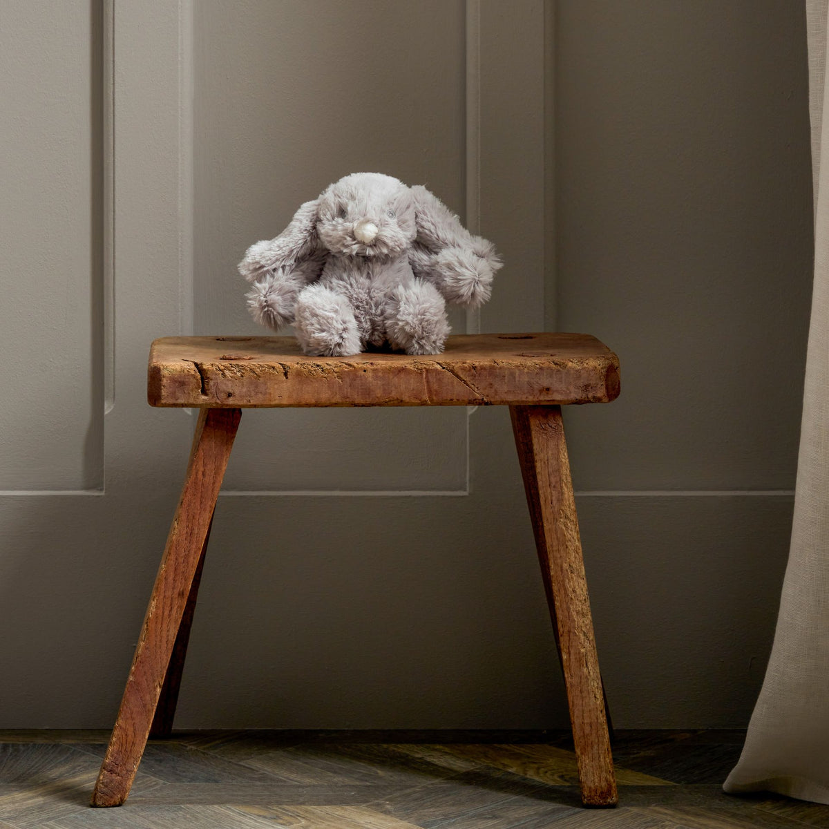 A small gray plush bunny toy sits on a rustic wooden stool in front of a gray paneled wall.