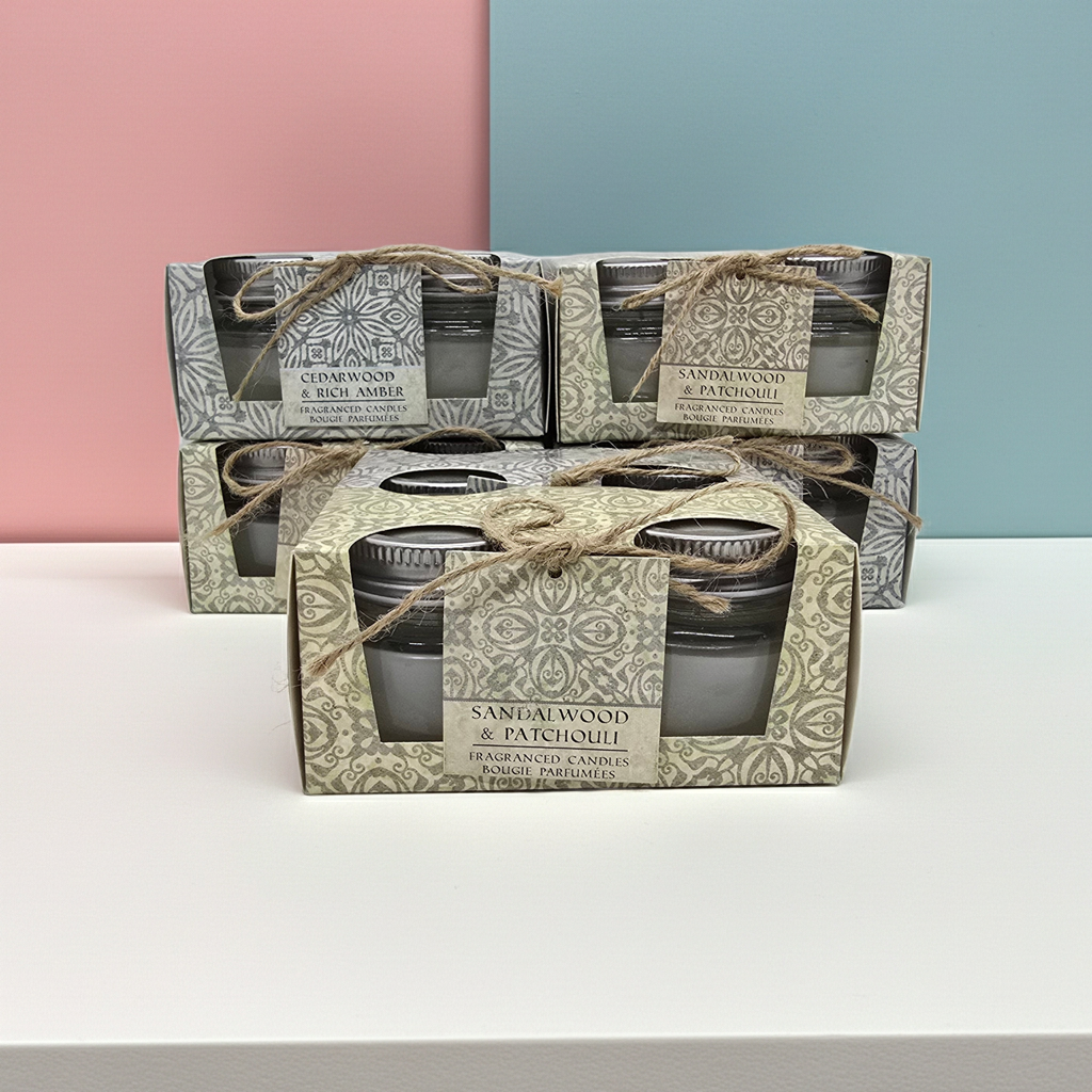 Stack of Scented candle boxes with 2 candles in glass jars per box 1 box has a Cedar wood and rich amber scent and the other has a sandalwood and patchouli scented candles. with decorative packaging on a pink and green gradient background.
