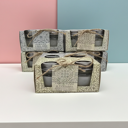Stack of Scented candle boxes with 2 candles in glass jars per box 1 box has a Cedar wood and rich amber scent and the other has a sandalwood and patchouli scented candles. with decorative packaging on a pink and green gradient background.