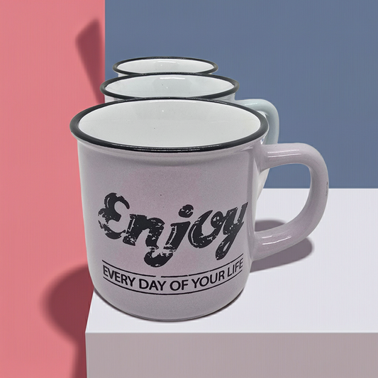 Pink mug with black text "Enjoy every day of your life" on a white surface against a pink and blue background