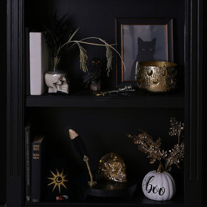 A dark, gothic-themed shelf display featuring a black owl figurine, a white ceramic skull with dried grass in a vase, a gold textured bowl, a framed black cat print, a black feather quill on a gold stand, a gold skull, and a white pumpkin with 'boo' written on it.