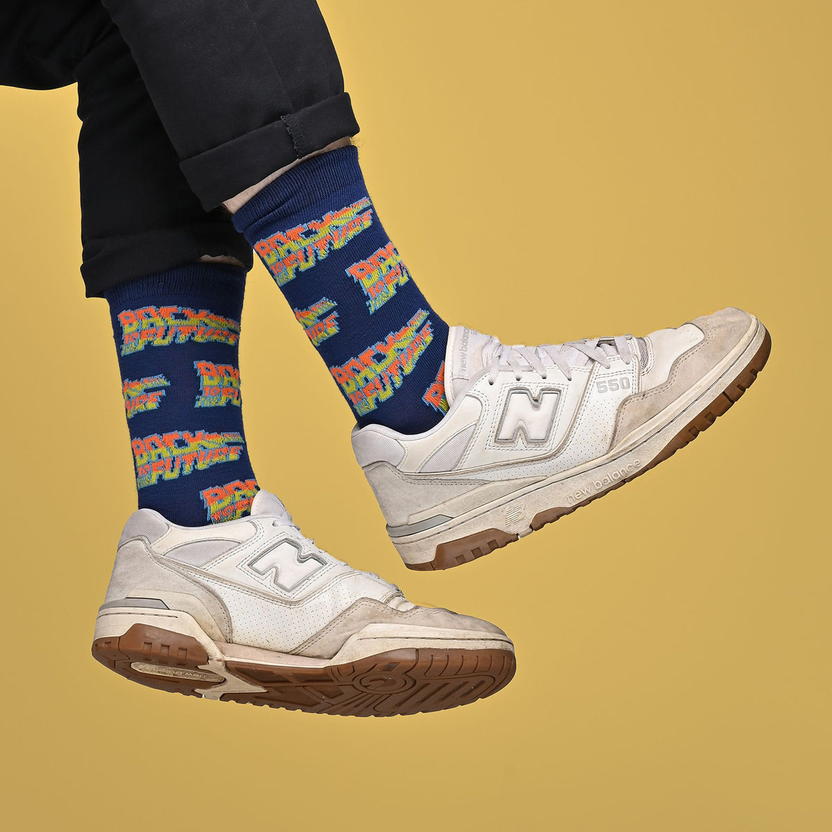 A person wearing navy blue socks with a colorful 'Back to the Future' pattern and white New Balance 990v2 sneakers, shown from the calves down against a solid mustard yellow background.