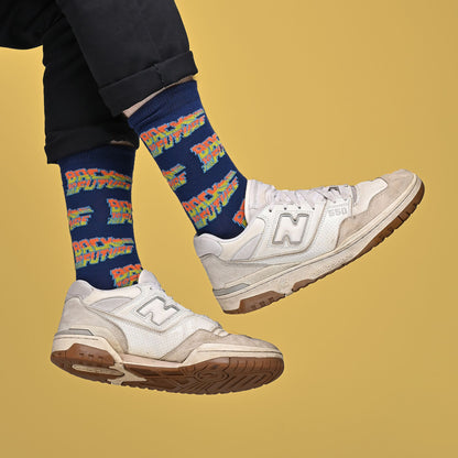 A person wearing navy blue socks with a colorful 'Back to the Future' pattern and white New Balance 990v2 sneakers, shown from the calves down against a solid mustard yellow background.