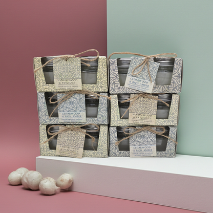 Stack of Scented candle boxes with 2 candles in glass jars per box 1 box has a Cedar wood and rich amber scent and the other has a sandalwood and patchouli scented candles. with decorative packaging on a pink and green gradient background.