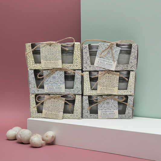 Stack of Scented candle boxes with 2 candles in glass jars per box 1 box has a Cedar wood and rich amber scent and the other has a sandalwood and patchouli scented candles. with decorative packaging on a pink and green gradient background.